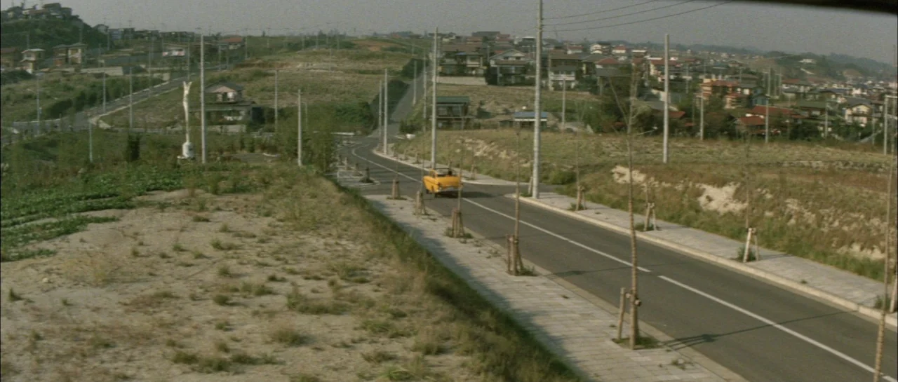 Godzilla Vs Megalon (1973) movie still: wide — A suburban landscape features rolling hills, sparse vegetation, and a winding asphalt road. A lone…