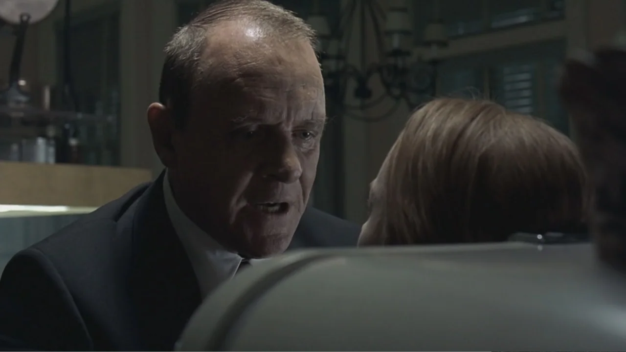 Hannibal (2001) movie still: medium — A dimly lit room with a muted color palette and soft shadow detail. An elderly man in a dark suit talks to an…