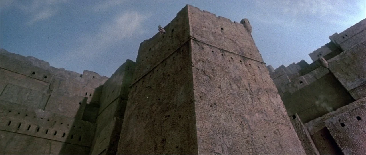 Never Say Never Again (1983) movie still: establishing — A massive, weathered stone fortress wall rises steeply against a pale, overcast sky. Small,…