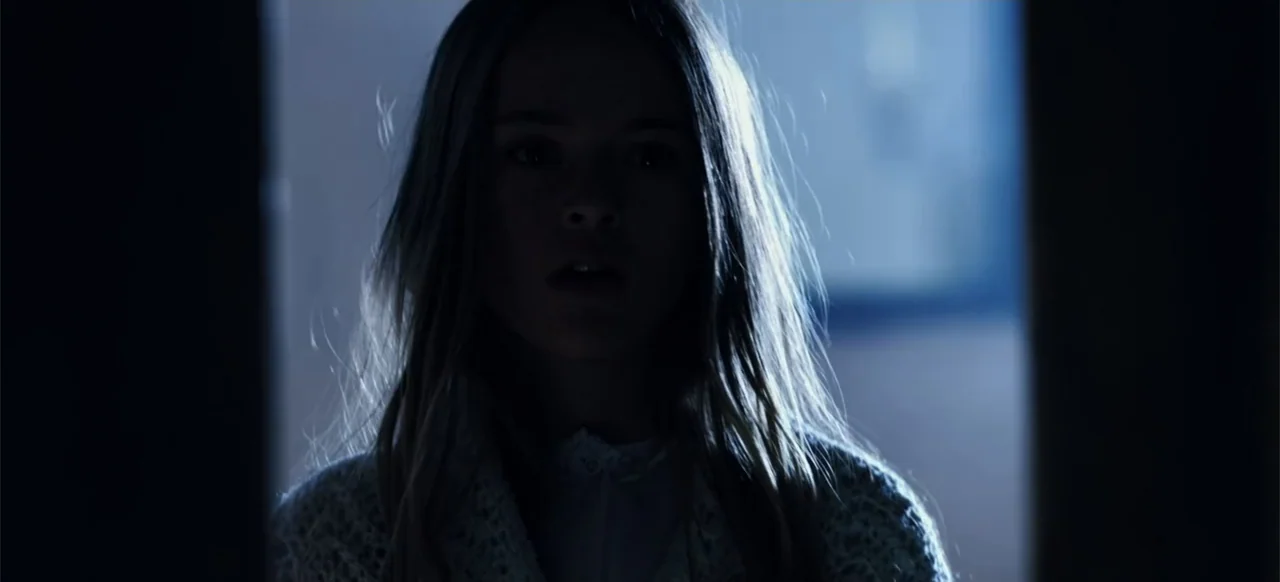 The Russian Bride (2019) movie still: closeup — A dark hallway transitions into a brightly lit space behind a young girl. The girl stands still in…