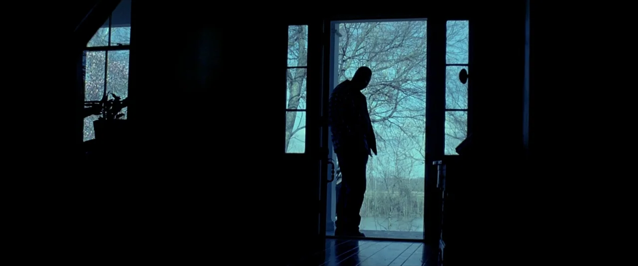 Ballast (2008) movie still: fullbody — A dimly lit interior room faces an open door showing a blue-toned exterior landscape. A man stands in the…