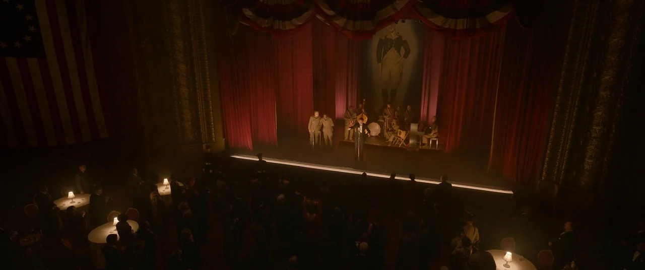Amsterdam (2022) movie still: wide — A dimly lit ballroom features a central stage with a red-curtained backdrop and musician. An audience sits at…
