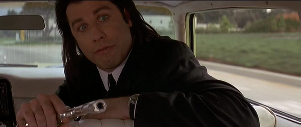 Pulp Fiction (1994) movie still: medium — A man in a black suit sits in the back of a moving vehicle. He holds a silver handgun while looking toward…
