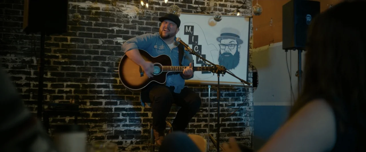 God Bless the Broken Road (2018) movie still: medium — A coffee shop interior features a dark painted brick wall adorned with light bulbs. A man…