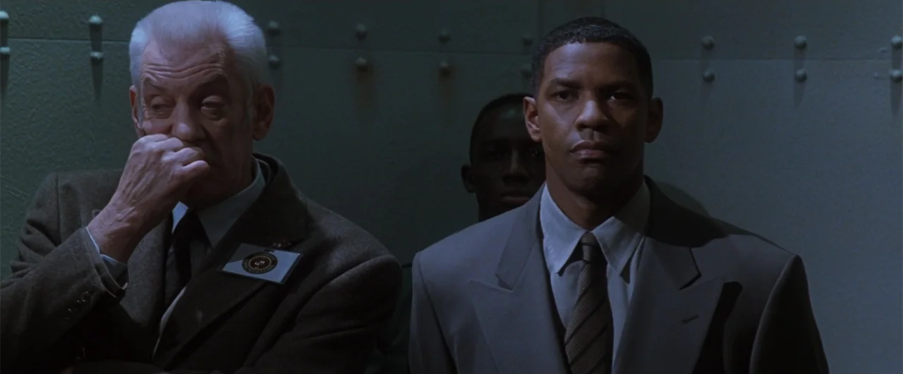 Fallen (2004) movie still: medium — Two men ride in a metallic elevator with large bolt heads visible on walls. The men stand side by side while a…