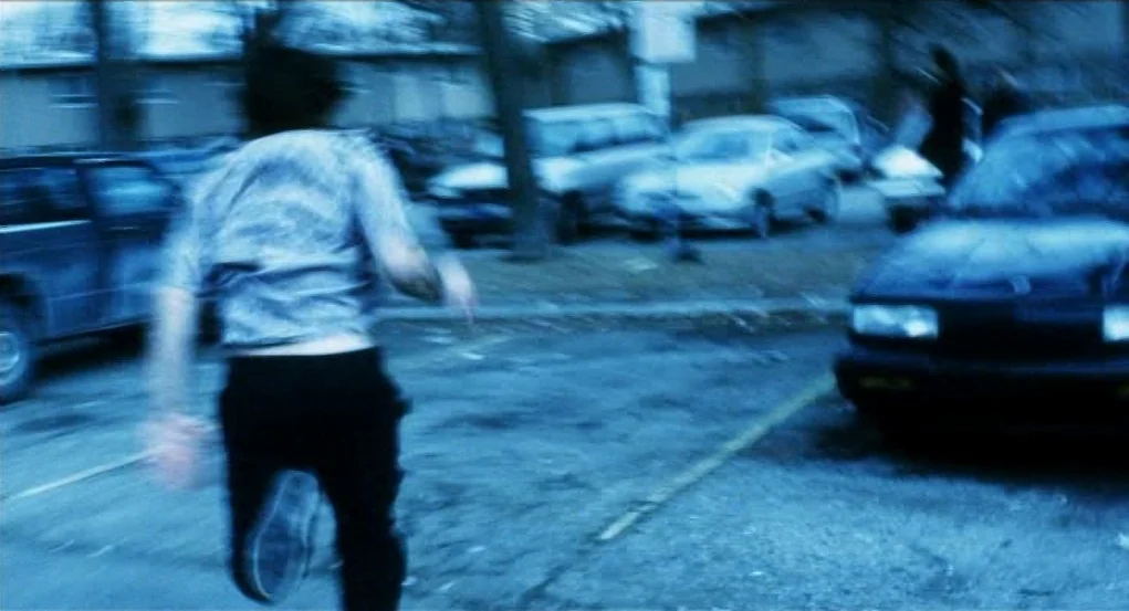 Narc (2002) movie still: fullbody — A blurred figure runs through a dark, paved outdoor area with multiple parked vehicles. The scene is enveloped…
