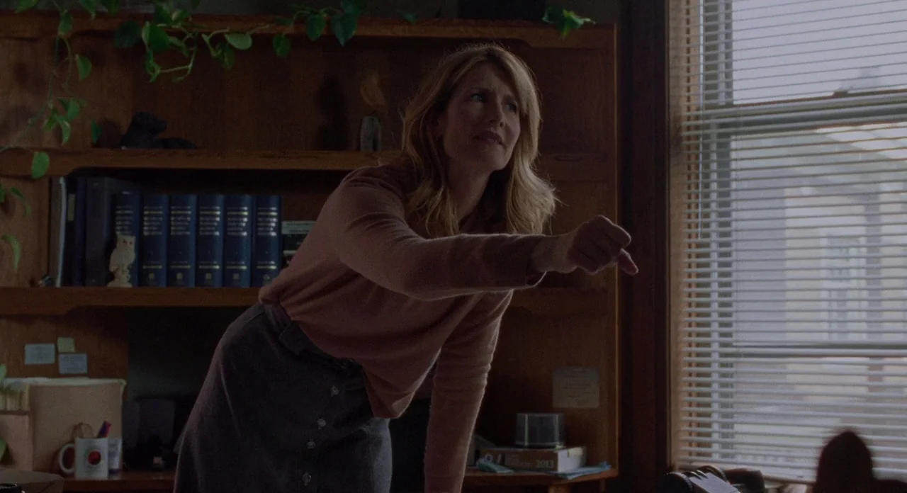 Certain Women (2016) movie still: medium — A woman leans over a desk in a room filled with wooden bookshelves and plants. She points her finger…