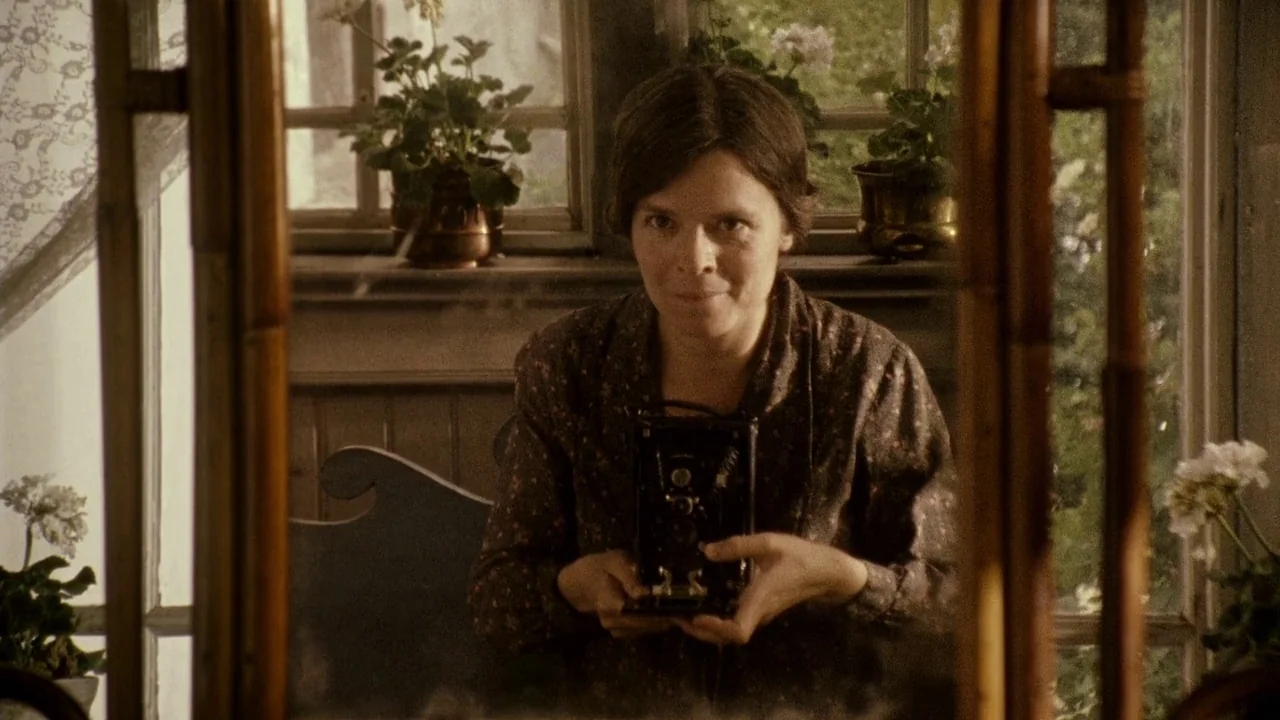 Everlasting Moments (2008) movie still: medium — A woman stands before a large, wood-framed mirror in a bright room with plants. She holds a vintage…
