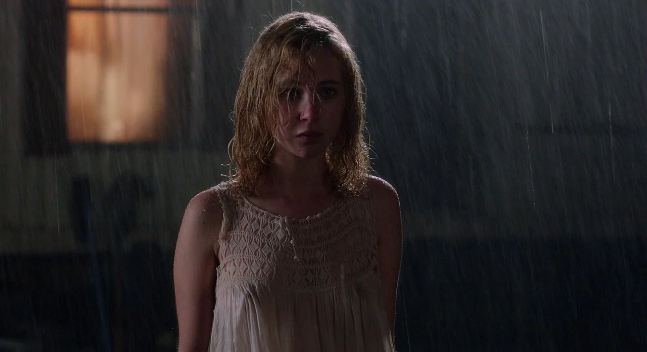Killer Joe (2011) movie still: medium — A young woman stands outdoors in heavy rain at night near a lighted window. She wears a light-colored dress…