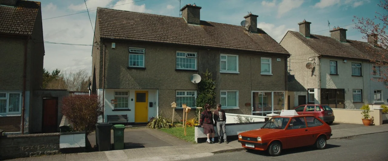 Extra Ordinary (2019) movie still: wide — A row of attached houses with grey stone facades faces a suburban paved street. Two people sit on a low…