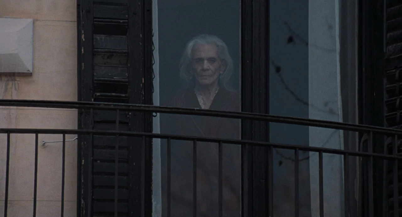 La Abuela (2021) movie still: medium — An elderly woman stands behind a glass pane overlooking an outdoor balcony. A dark metal railing occupies the…