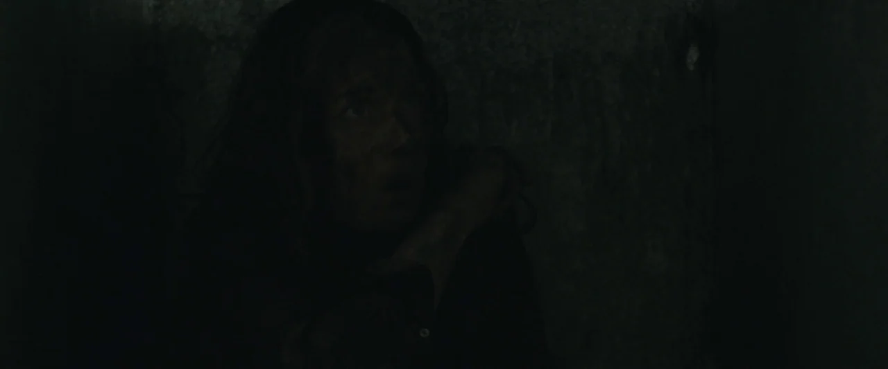 Here Alone movie still: closeup — A very dark, nearly black space with indistinct textures on the surrounding walls. A woman with smudged cheeks…