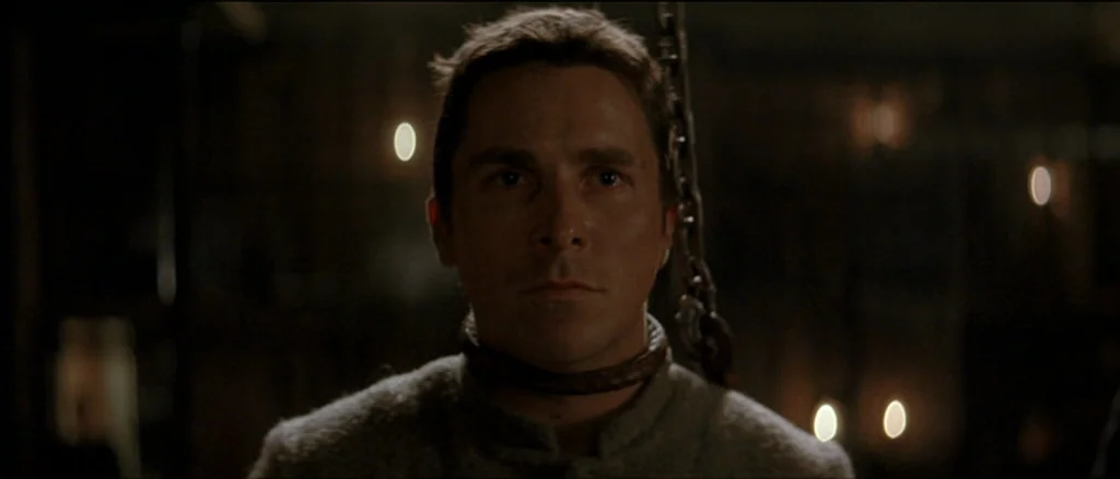 The Prestige (2006) movie still: closeup — A dimly lit, stone-walled dungeon interior features a man wearing an iron collar. The man looks forward,…