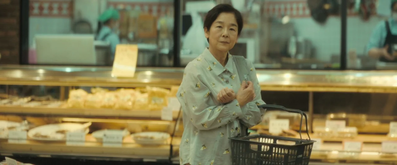 Monster (2023) movie still: medium — An elderly woman stands before a display case filled with baked goods. She holds a black plastic shopping…
