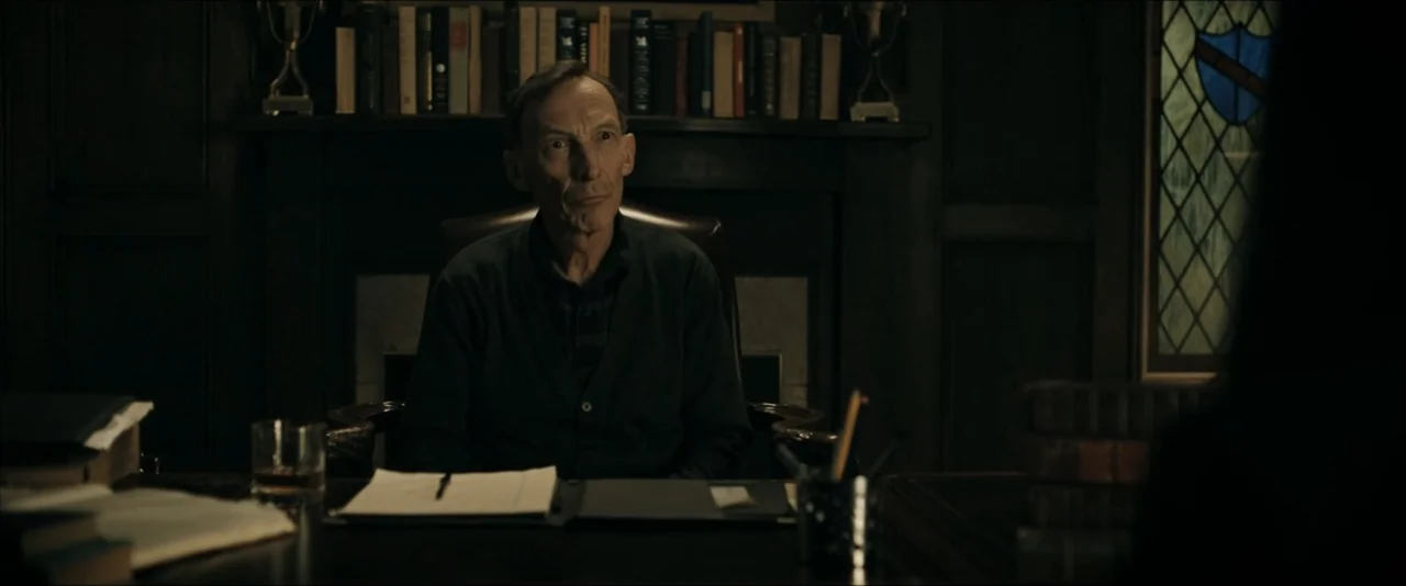 It Feeds (2025) movie still: medium — A dark, wood-paneled study features a desk filled with papers and books. An elderly man sits in a leather…