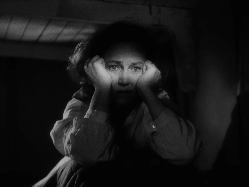 A Ship Bound for India (1947) movie still: closeup — A dark, cramped interior with metallic rivets visible on the overhead structure. A woman sits…