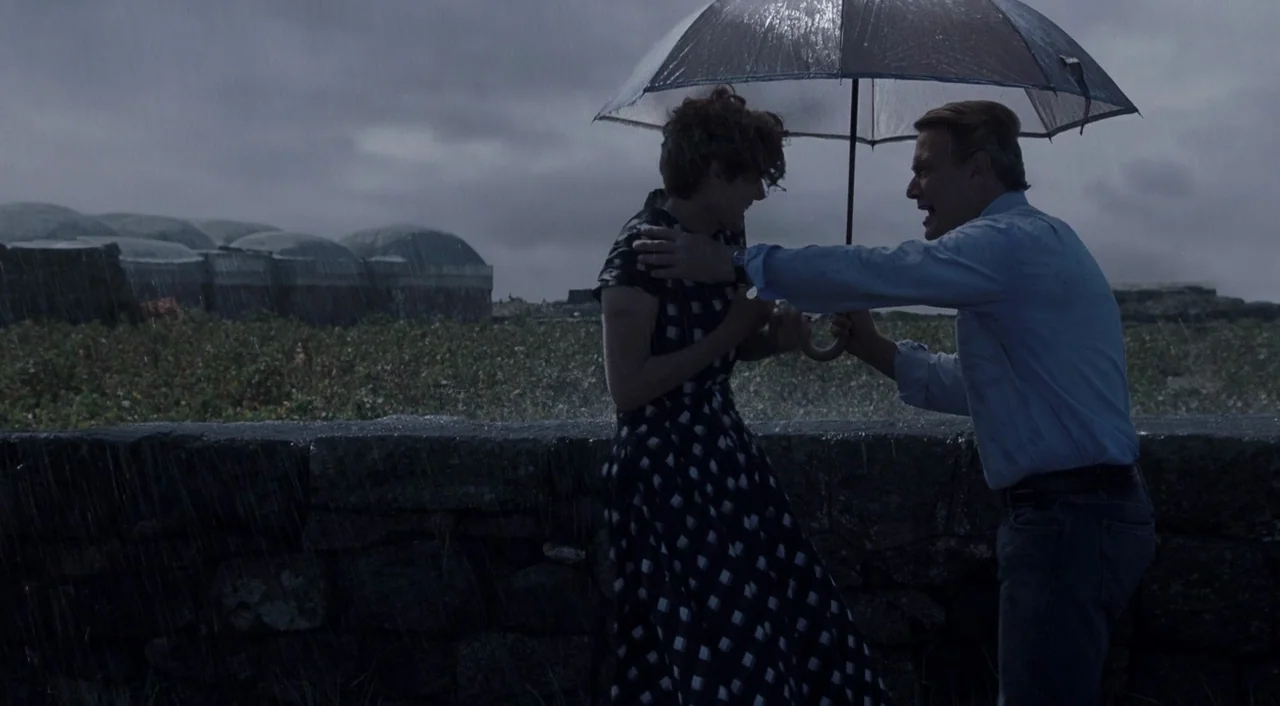 A Bigger Splash (2015) movie still: fullbody — An open coastal landscape features a stone wall under a heavy, overcast sky. A man holds an umbrella…
