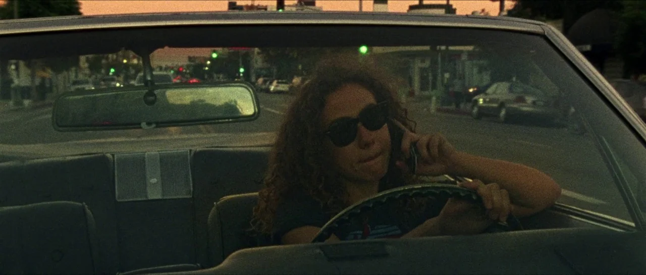 Bliss movie still: medium — A woman drives a car along a city street during the evening. She holds a smartphone to her ear while steering through…