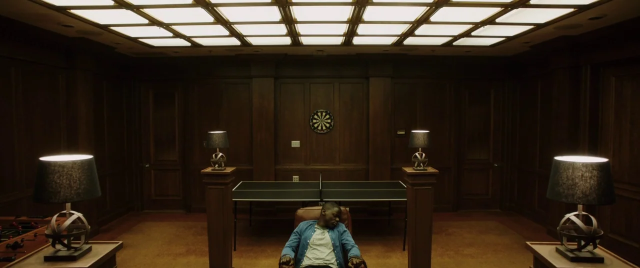 Get Out (2017) movie still: wide — A symmetrical room finished with dark wood paneling features a centered ping pong table. A lone man sits in a…