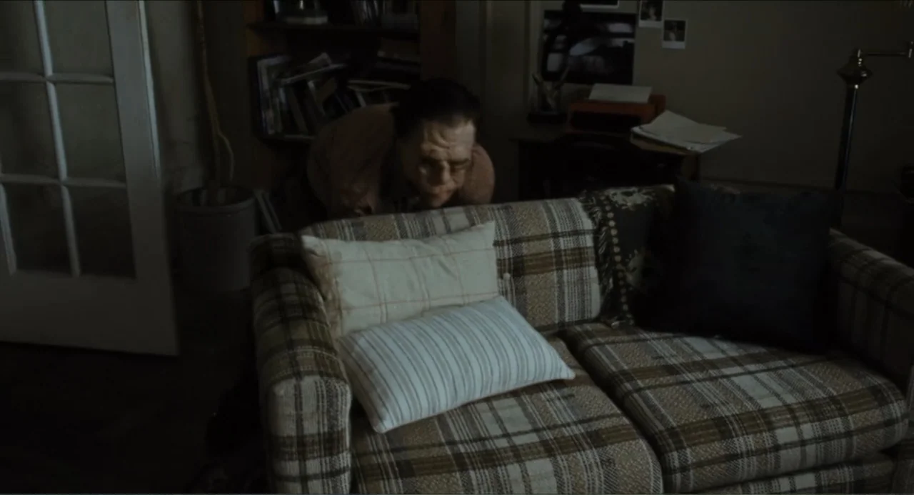 A Different Man (2024) movie still: medium — A dimly lit room features a plaid sofa set against a dark wall. A man leans over the back of the couch…
