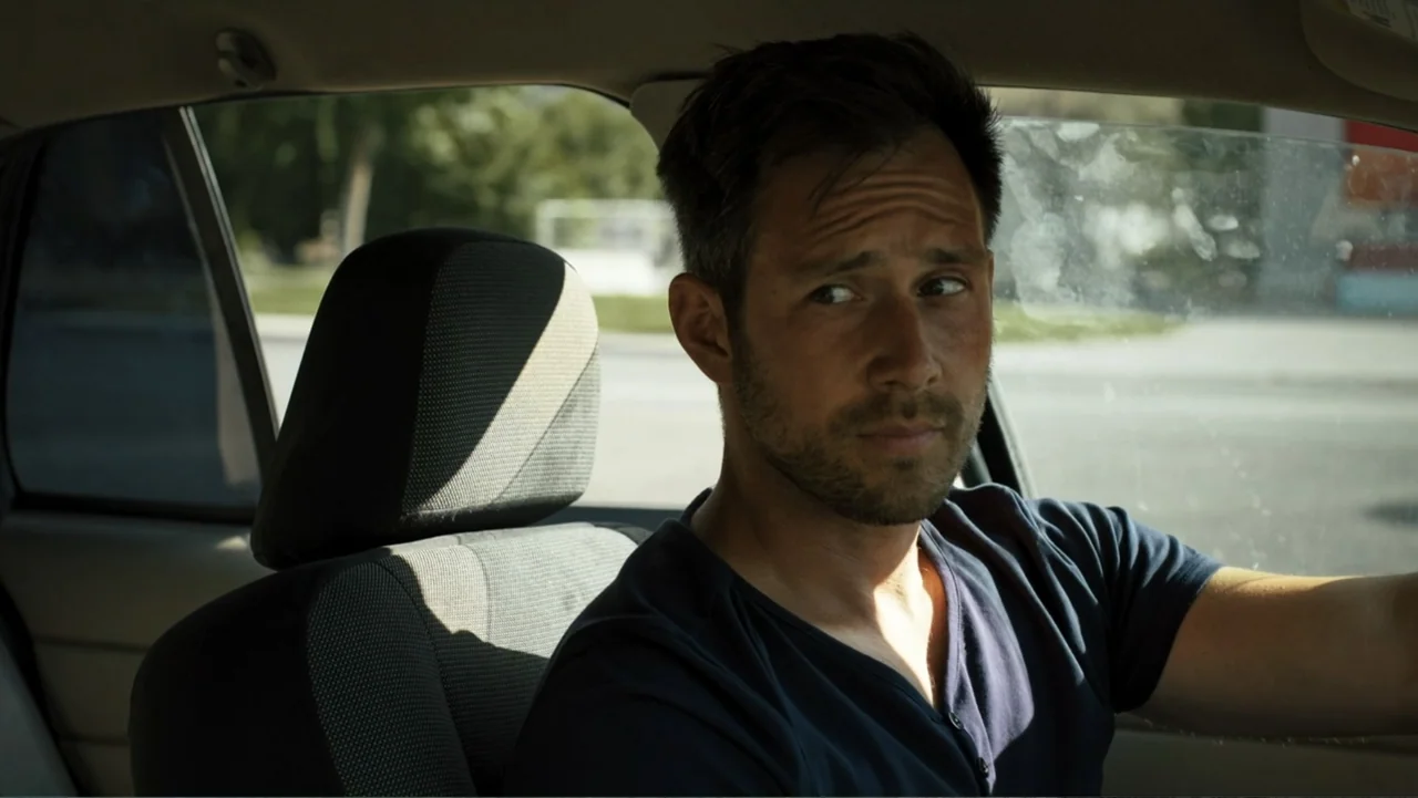 Bullet Proof (2022) movie still: medium — A man sits in the driver seat of a car parked during the day. He wears a navy blue shirt and looks toward…
