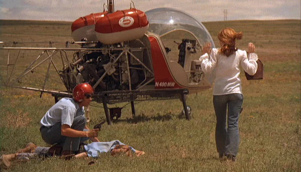 Badlands (1973) movie still: fullbody — An open grassy field contains a small red and white helicopter parked on the ground. A man crouches with a…
