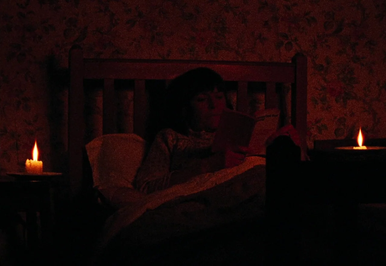 Enys Men (2022) movie still: medium — A dimly lit room features patterned wallpaper and a dark wooden bed headboard. A woman sits propped up in bed,…