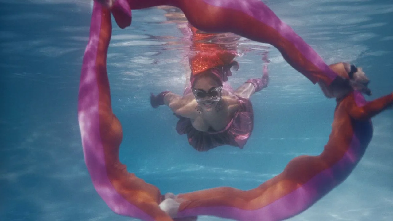 Black is King (2020) movie still: medium — A person swims underwater in a bright blue pool. They pass through a circular fabric ring.