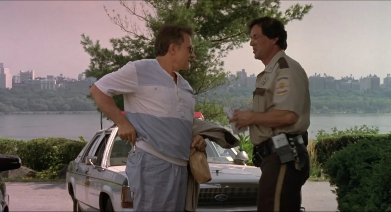 Cop Land (1997) movie still: medium — A riverside park area with a city skyline visible across a wide body of water. A man in civilian clothes…
