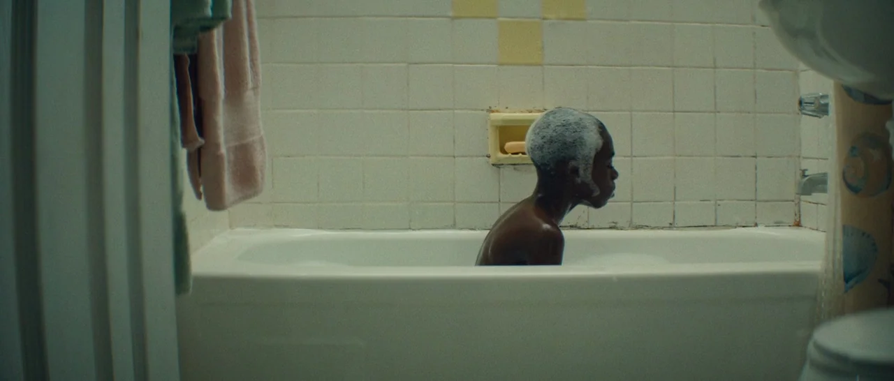 Moonlight (2016) movie still: medium — A bathroom features white tiled walls with a built-in yellow recessed soap dish. A young boy sits in a white…
