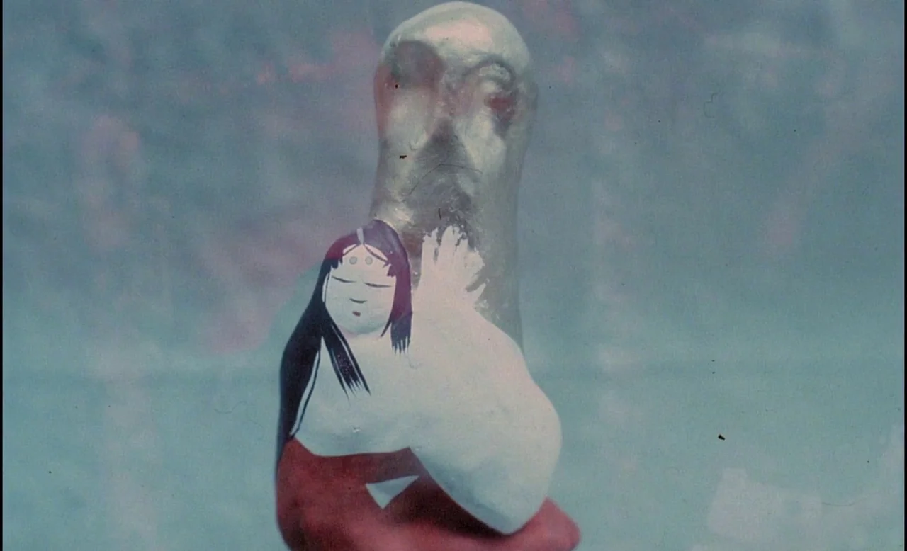 Sans Soleil (1983) movie still: closeup — A small, painted figurine of a woman rests against a smooth, metallic, upright sculpture. The background…