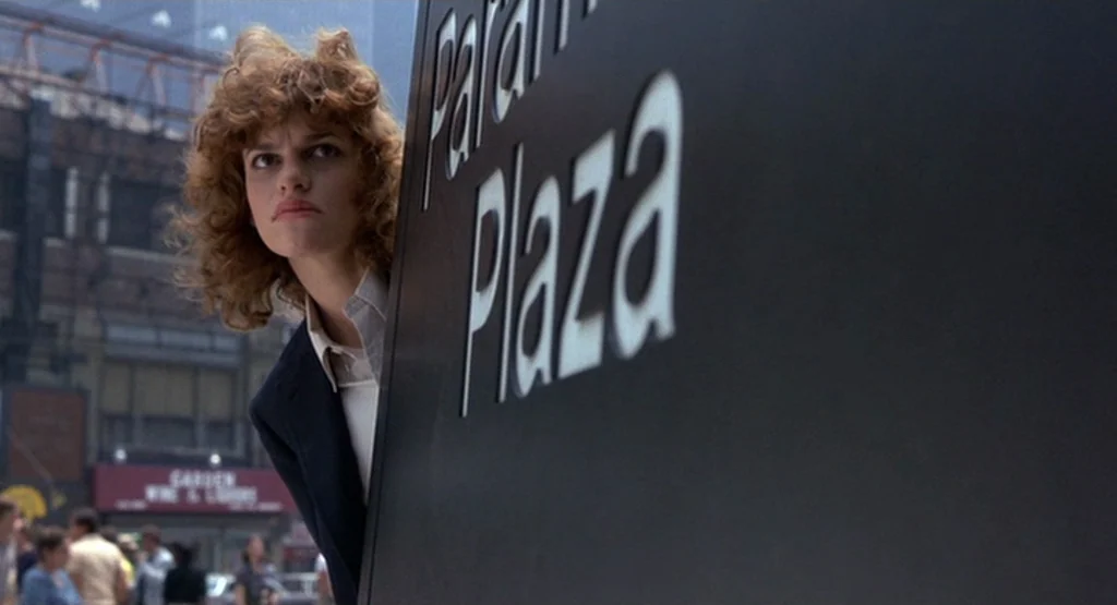 The King Of Comedy (1982) movie still: medium — A woman stands behind a large black sign labeled Param Plaza on a street. She peers around the…