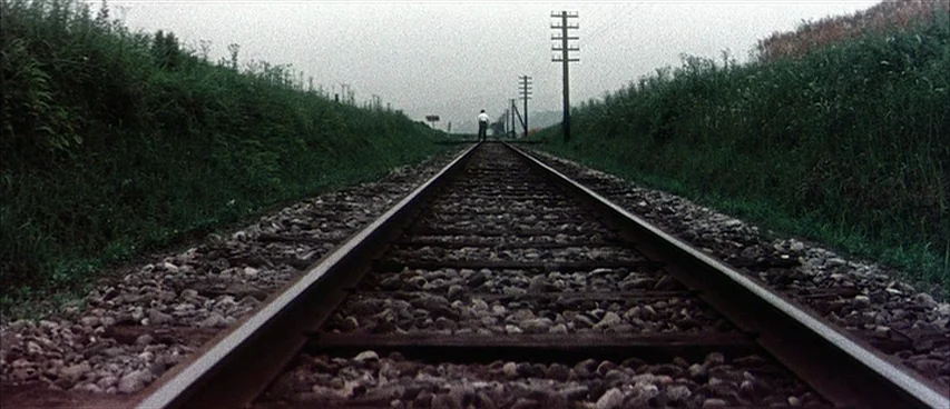 Jigoku (1960) movie still: wide — A long stretch of railway tracks cuts through a grassy corridor under overcast skies. A lone figure stands on the…