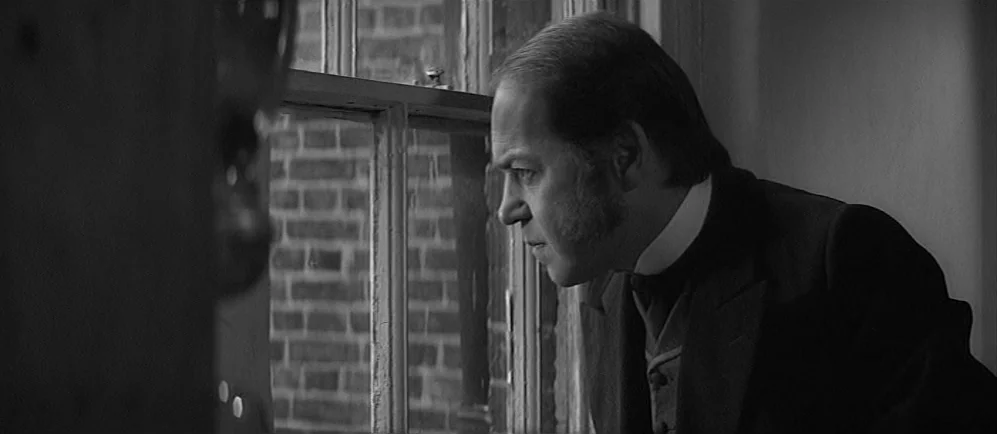 The Elephant Man (1980) movie still: medium — An interior space features a wooden-framed window overlooking a brick building facade. A man in a…