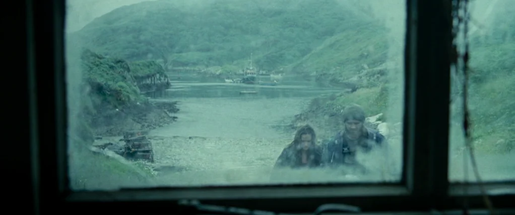 Ondine (2009) movie still: wide — A desolate coastal inlet is visible through a dirty, fogged-up window frame. A man and a woman walk along the…