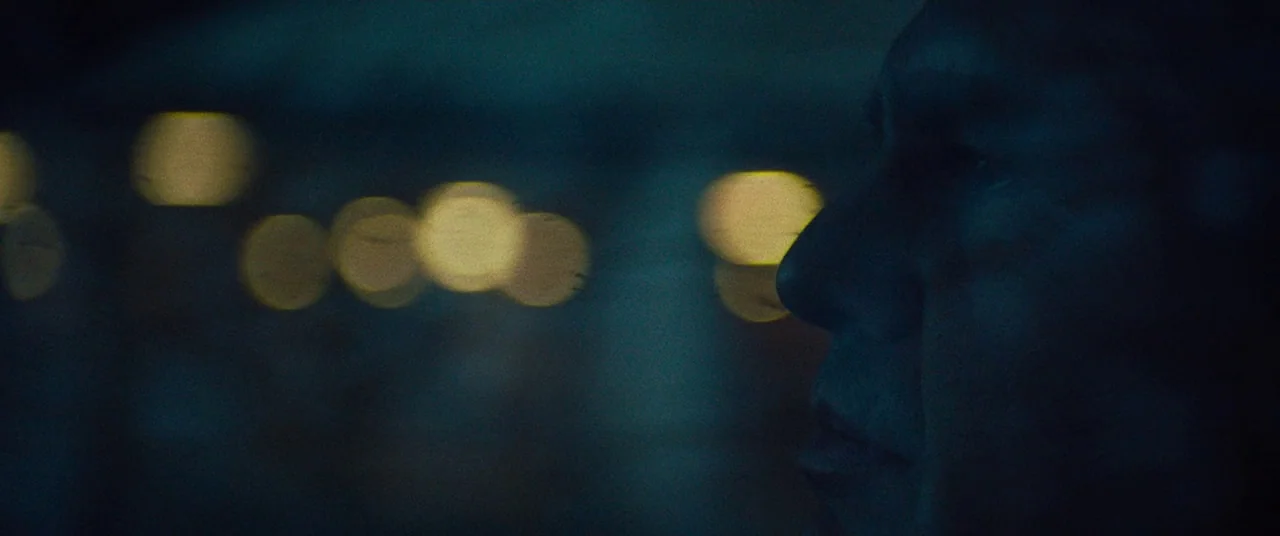 Dark Waters (2019) movie still: closeup — A dark, out-of-focus view of city lights at night viewed through a window. A man's face is seen in profile…