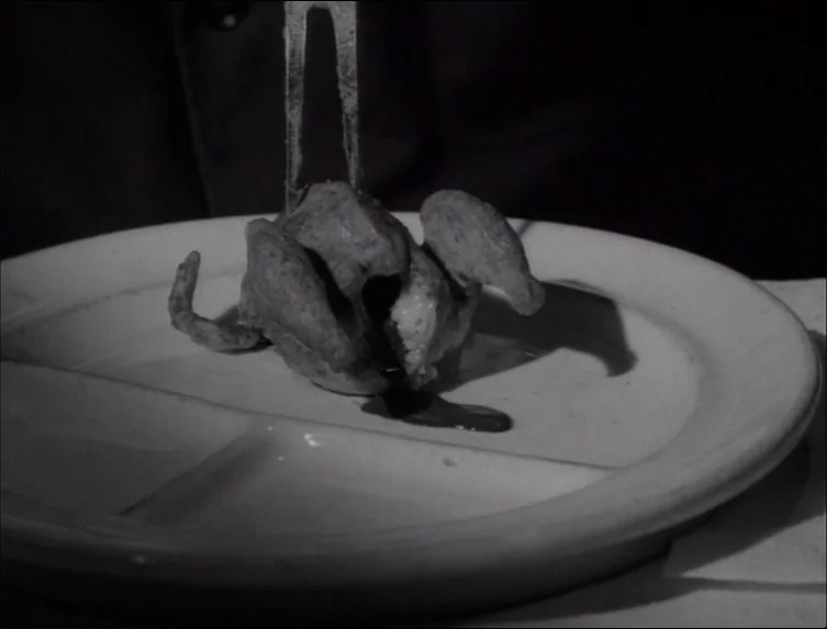 Eraserhead (1977) movie still: closeup — A close up view of a small roasted bird on a white partitioned plate. A silver fork presses into the center…
