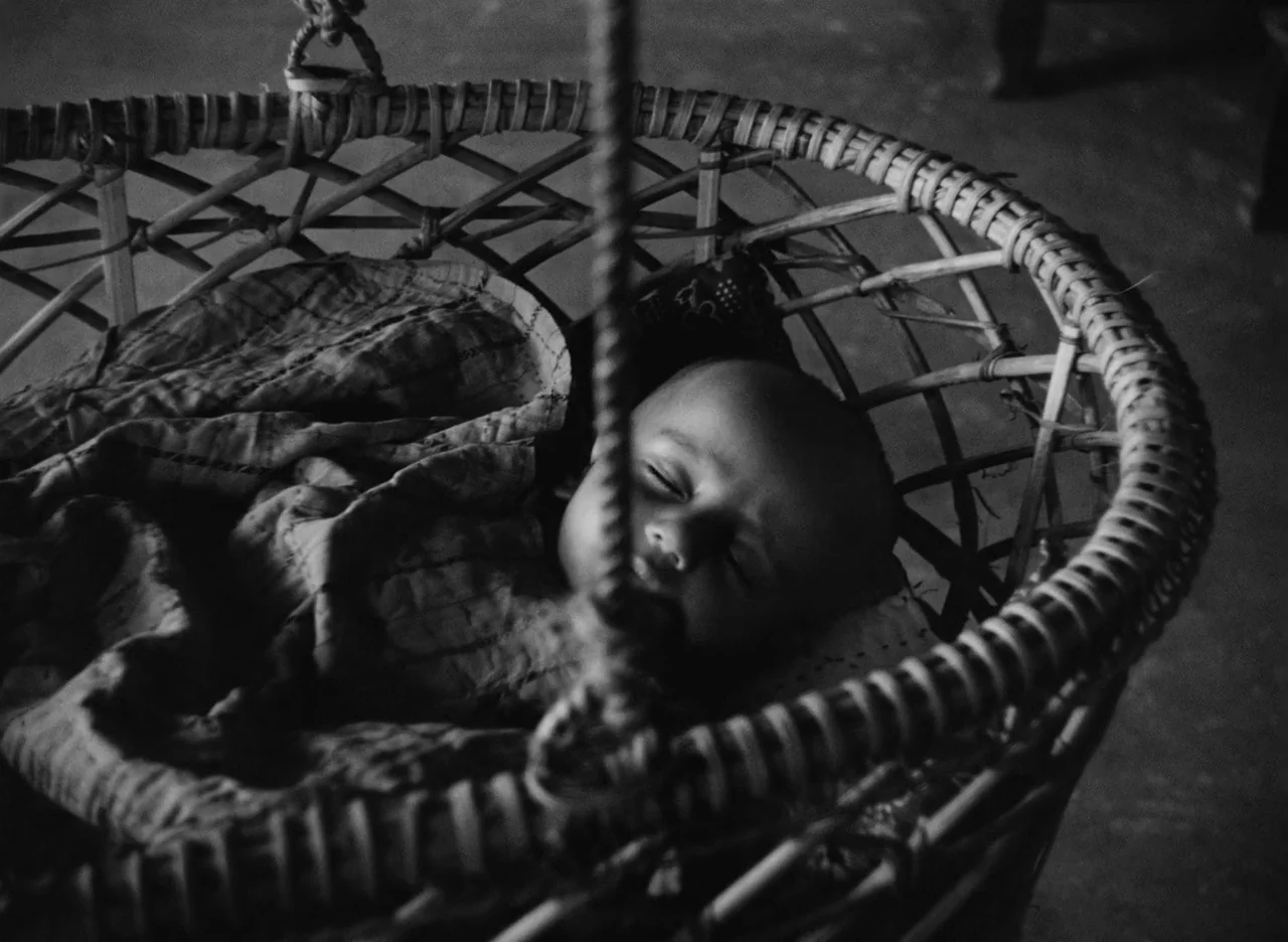 Apur Sansar (1959) movie still: closeup — A handmade woven wicker cradle hangs in a dim room with rustic textures. A small infant rests sleeping…