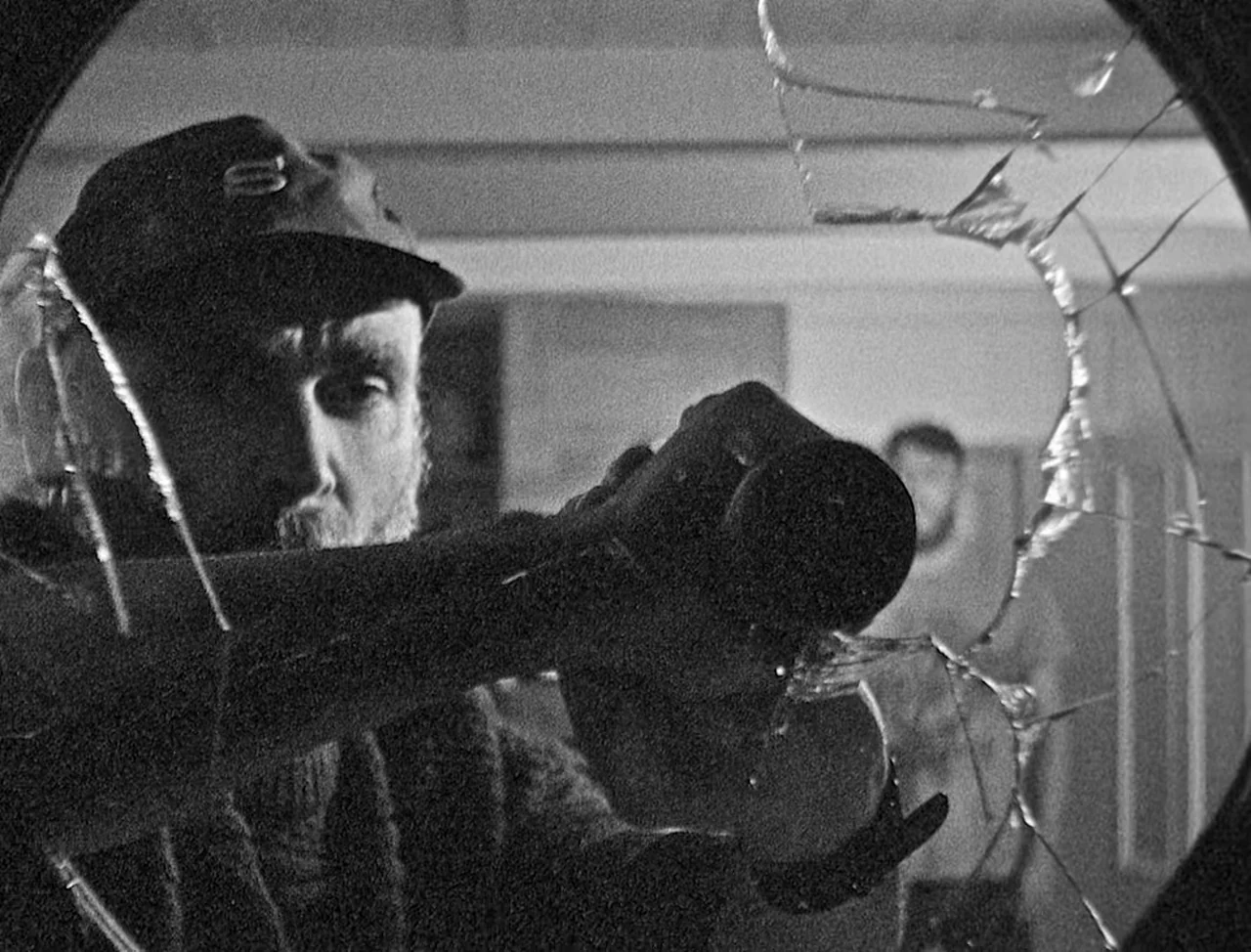 Bait (2019) movie still: closeup — A man holds a camera behind a pane of shattered, circular-framed glass. The subject looks through the lens while…