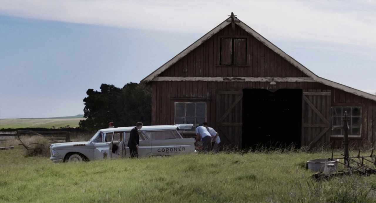 X (2022) movie still: wide — An old red wooden barn stands in a grassy field on a clear day. Three men in professional attire congregate around the…