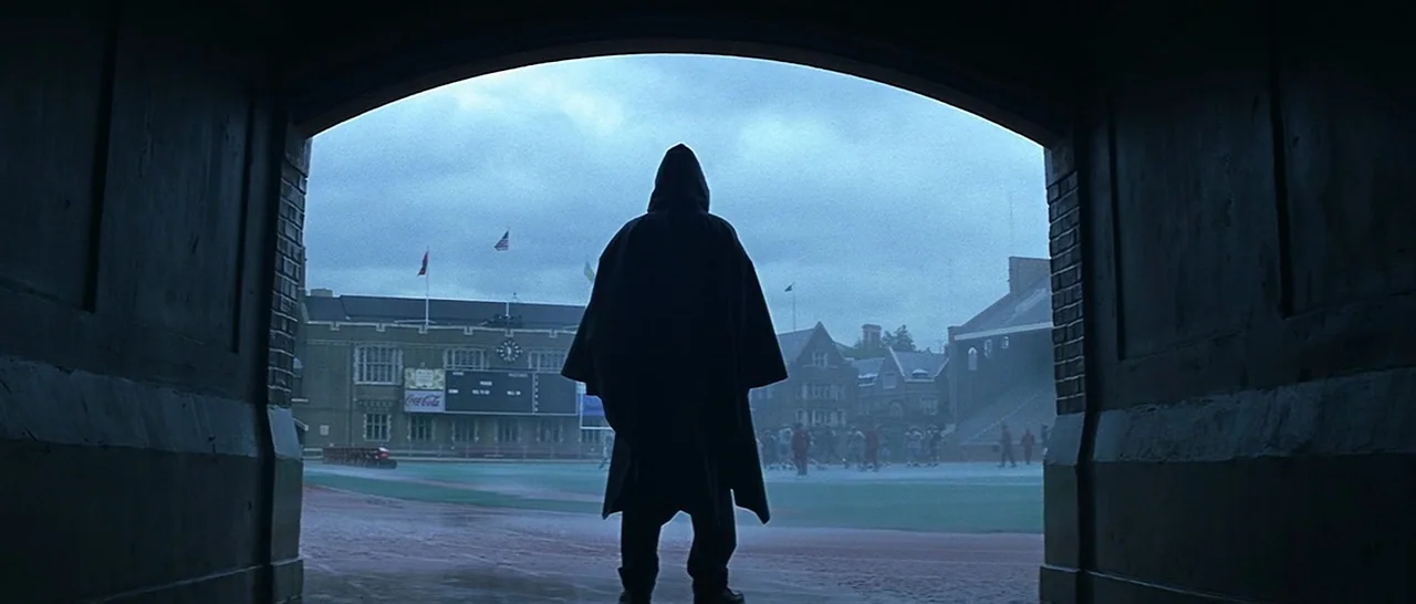 Unbreakable (2000) movie still: fullbody — A dark brick tunnel opens out onto a green athletic field under overcast skies. A figure in a black…