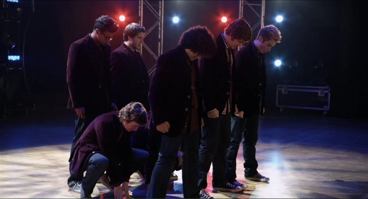 Pitch Perfect (2012) movie still: fullbody — A dark wooden stage illuminated by colored spotlights with metal trussing in the background. Five young…