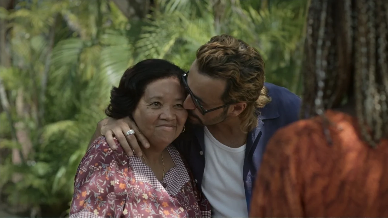 Unknown #1 movie still: medium — A lush tropical garden provides a dense green background of palm fronds and leaves. A younger man embraces and…