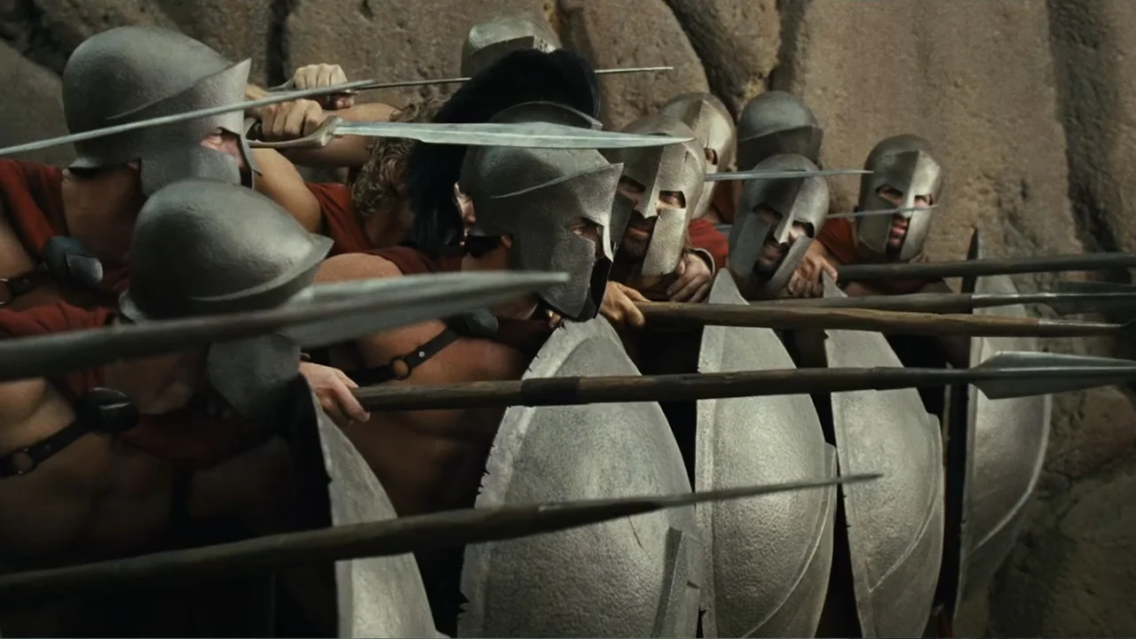 Meet the Spartans (2008) movie still: medium — A phalanx of Spartan soldiers stands against a textured cliff face in a mountain pass. The men clutch…