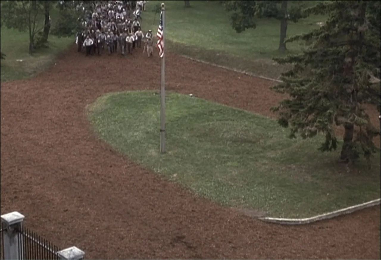 Vendetta (2022) movie still: wide — A wide outdoor scene features a large group walking across a brown dirt path. A central flagpole with a United…