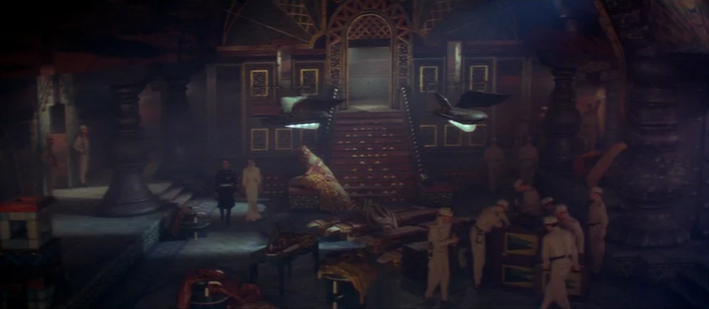 Dune (1984) movie still: wide — A vast, dimly lit hall features ornate, dark-toned architectural pillars and a central staircase. Several figures in…