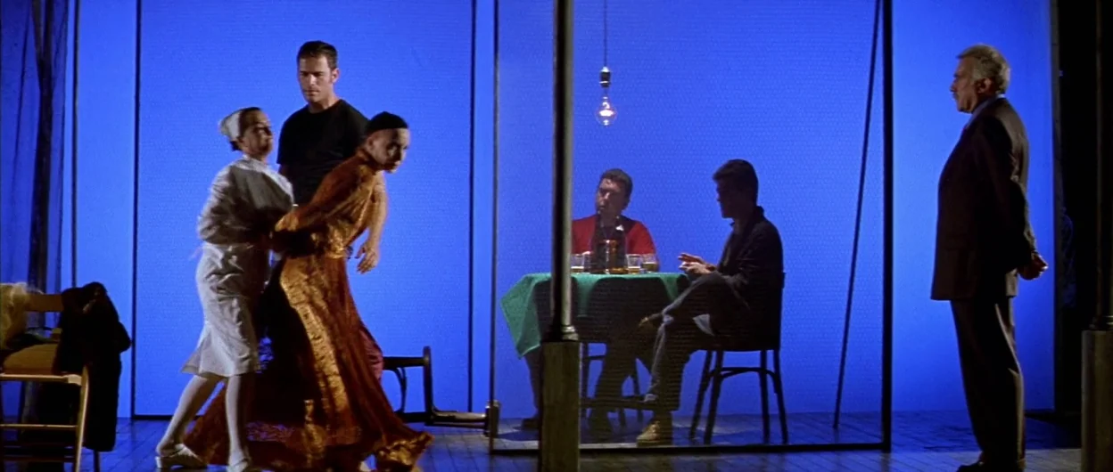 All About My Mother (1999) movie still: wide — A theatrical stage set features a vibrant blue backdrop behind a simple table arrangement. Six actors…
