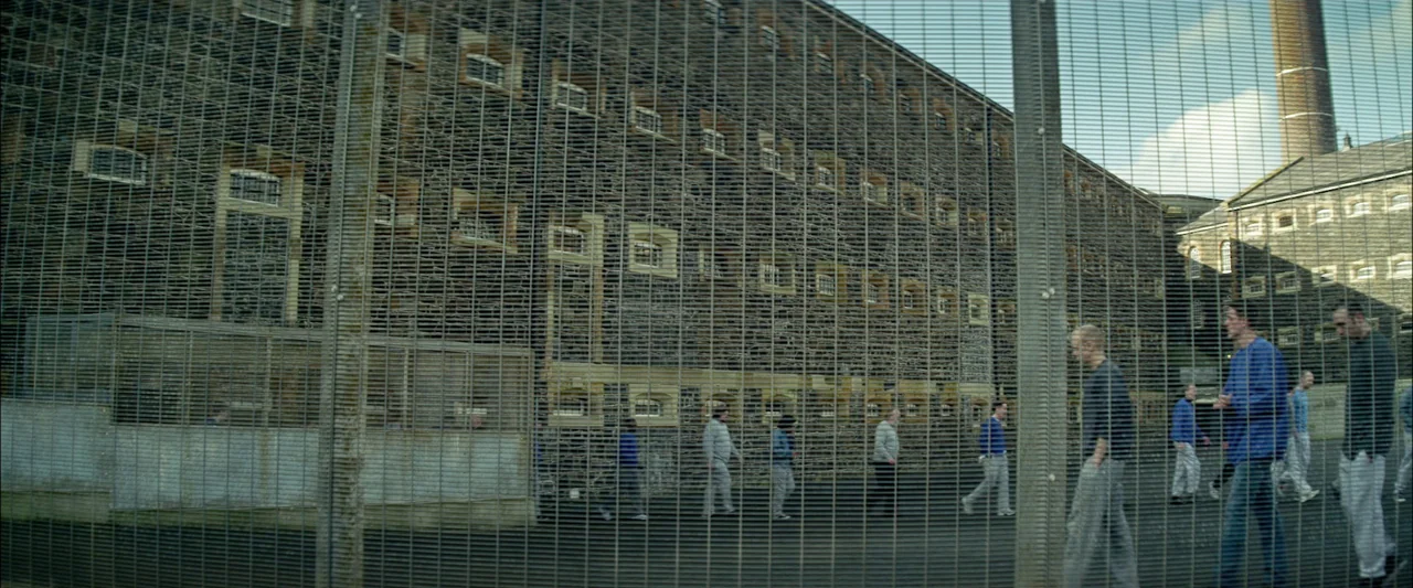 Starred Up (2013) movie still: wide — A large multi-story stone building sits behind a high heavy-duty chain-link security fence. A group of men in…