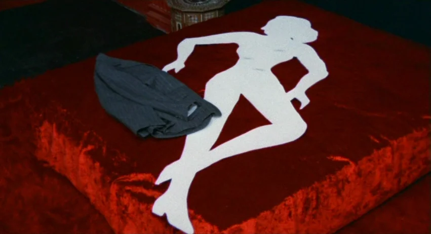 A Lizard In A Woman’s Skin (1971) movie still: medium — A low-profile surface covered in dark red velvet textured fabric sits in a room. A white…