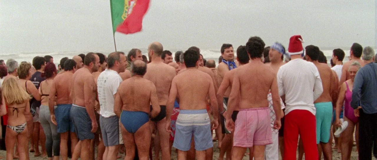 Arabian Nights: Volume 1 – The Restless One (2015) movie still: wide — A crowd of people stands on a sandy beach under an overcast grey sky. A large…